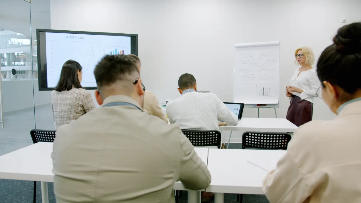 Equipe reunida em sala de treinamento analisando gráficos e relatórios em monitores e quadros, enquanto uma instrutora apresenta conteúdo para participantes sentados e em pé.