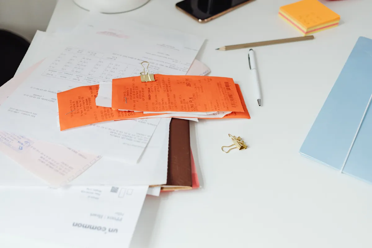 Mesa de escritório com documentos e folhas com anotações, clipes, marcador de papel em laranja e lápis sobre base branca, sugerindo organização e preparação para reunião ou trabalh