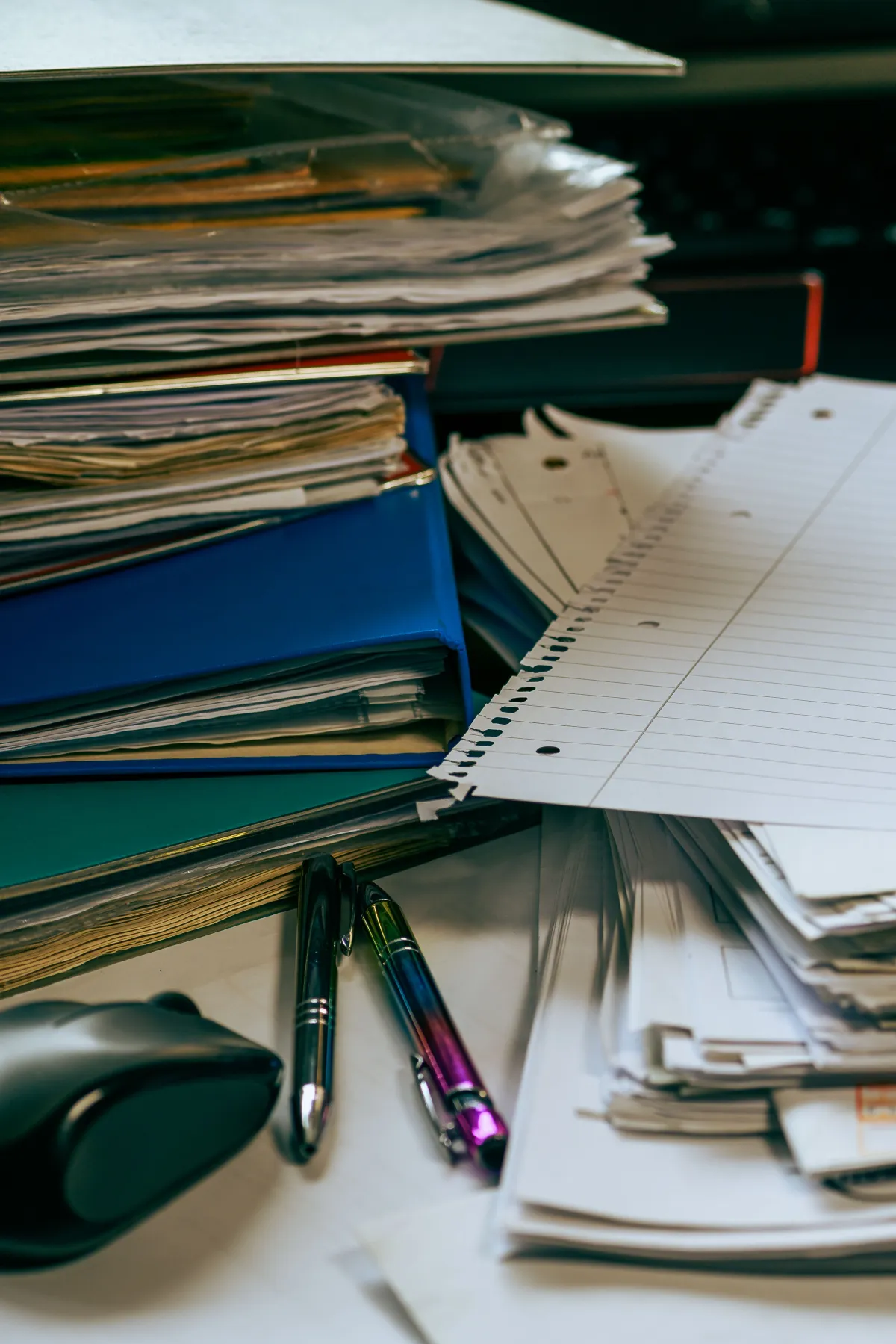 Pilhas de cadernos e folhas com anotações, uma agenda aberta com páginas pautadas e canetas sobre a mesa, sugerindo estudo e organização
