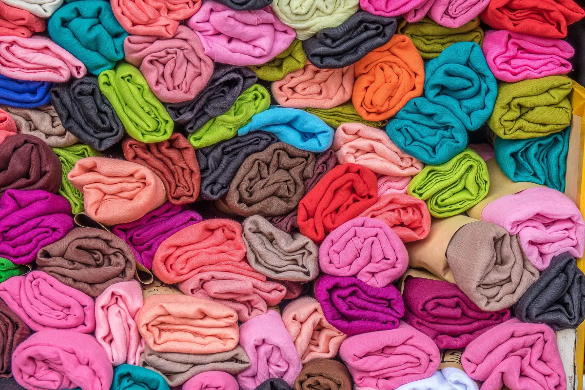 Fundo com várias peças de tecido dobradas em cores vibrantes, incluindo rosa, verde, azul, laranja, vermelho e roxo, formando um mosaico têxtil.