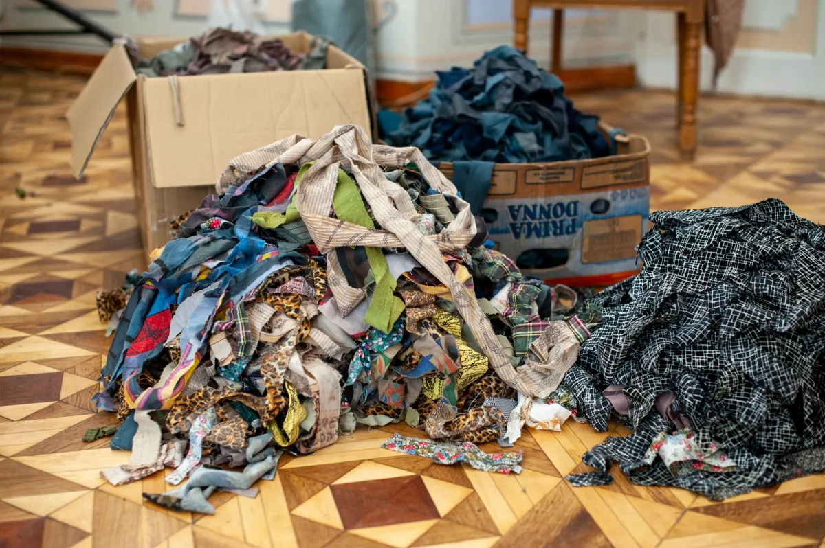 Pilha de roupas e tecidos misturados em caixas e no chão, com peças coloridas e estampadas em uma sala de estar, sugerindo separação ou doação de vestuário.