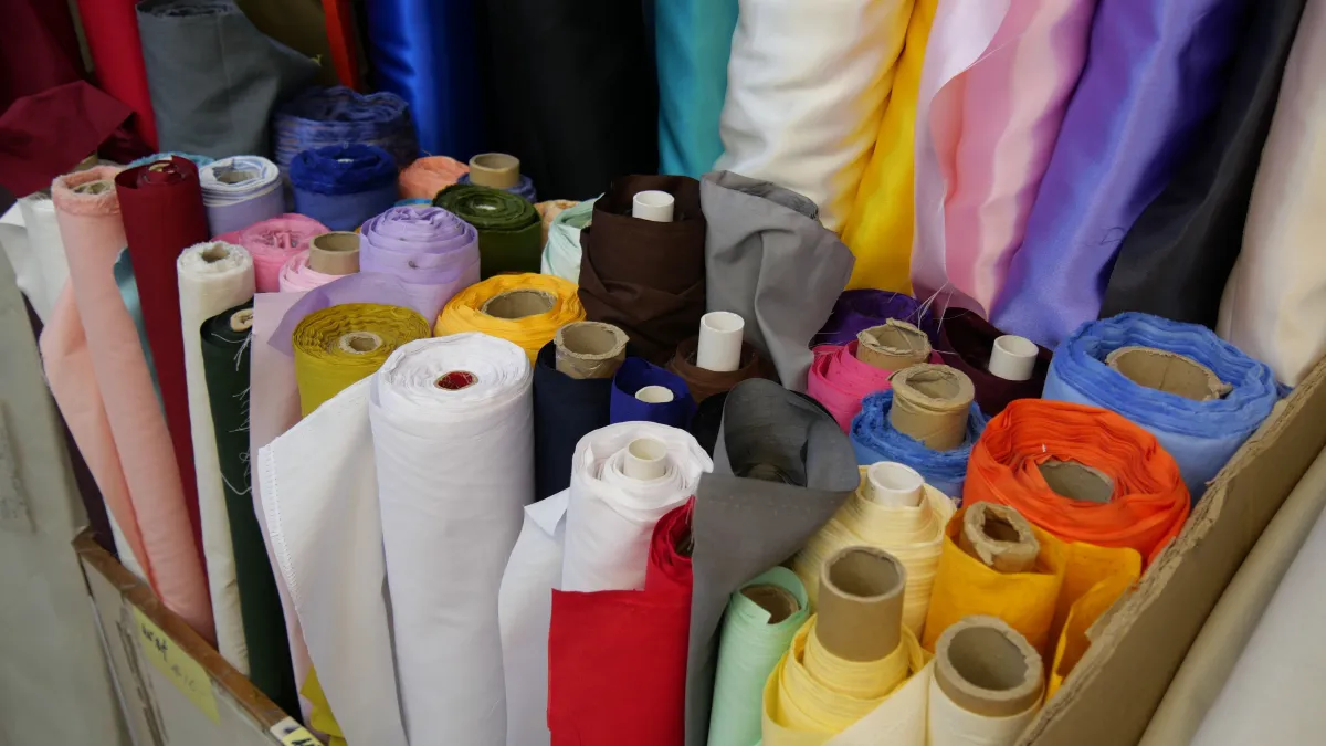 Caixa com rolos de tecidos coloridos e estampados, incluindo algodão, linho e materiais em tons como branco, vermelho, azul, amarelo e roxo, prontos para confecção.