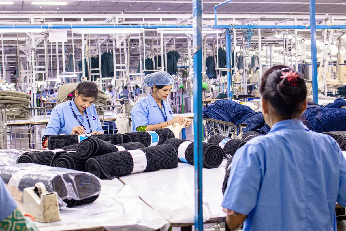 Funcionárias em uma fábrica têxtil inspecionando e organizando rolos de tecidos escuros em uma bancada, com equipamentos e estruturas industriais ao fundo.