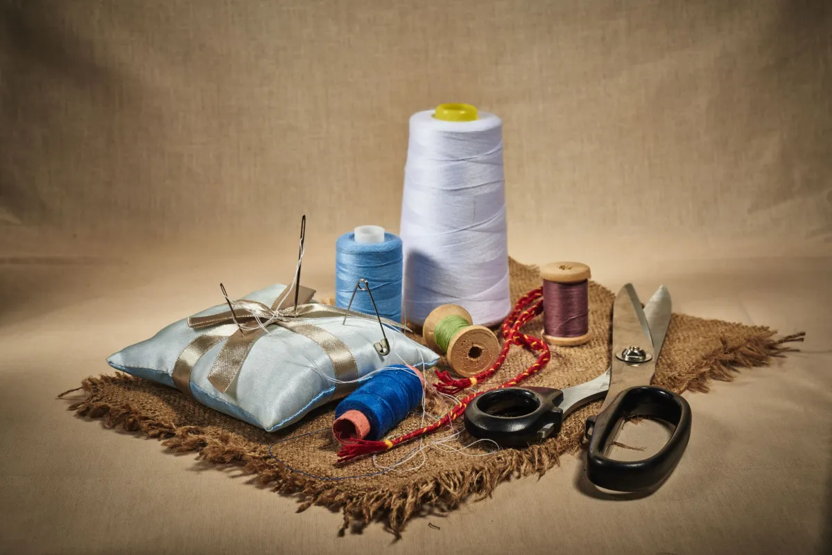 Conjunto de materiais de costura em fundo neutro: carretéis de linha branca e azul, tesoura, alfinetes, fita, peça de tecido e linha colorida para artesanato e reparos