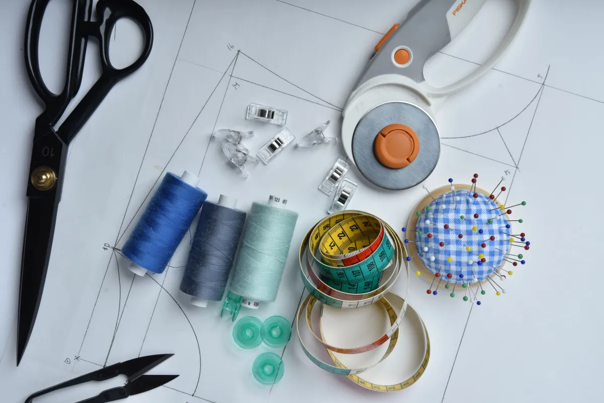 Materiais de costura e artesanato sobre um molde de modelagem: tesoura, carretéis de linha azul, verde e cinza, fita métrica, rolo de fita e alfineteiro com alfinhos coloridos.