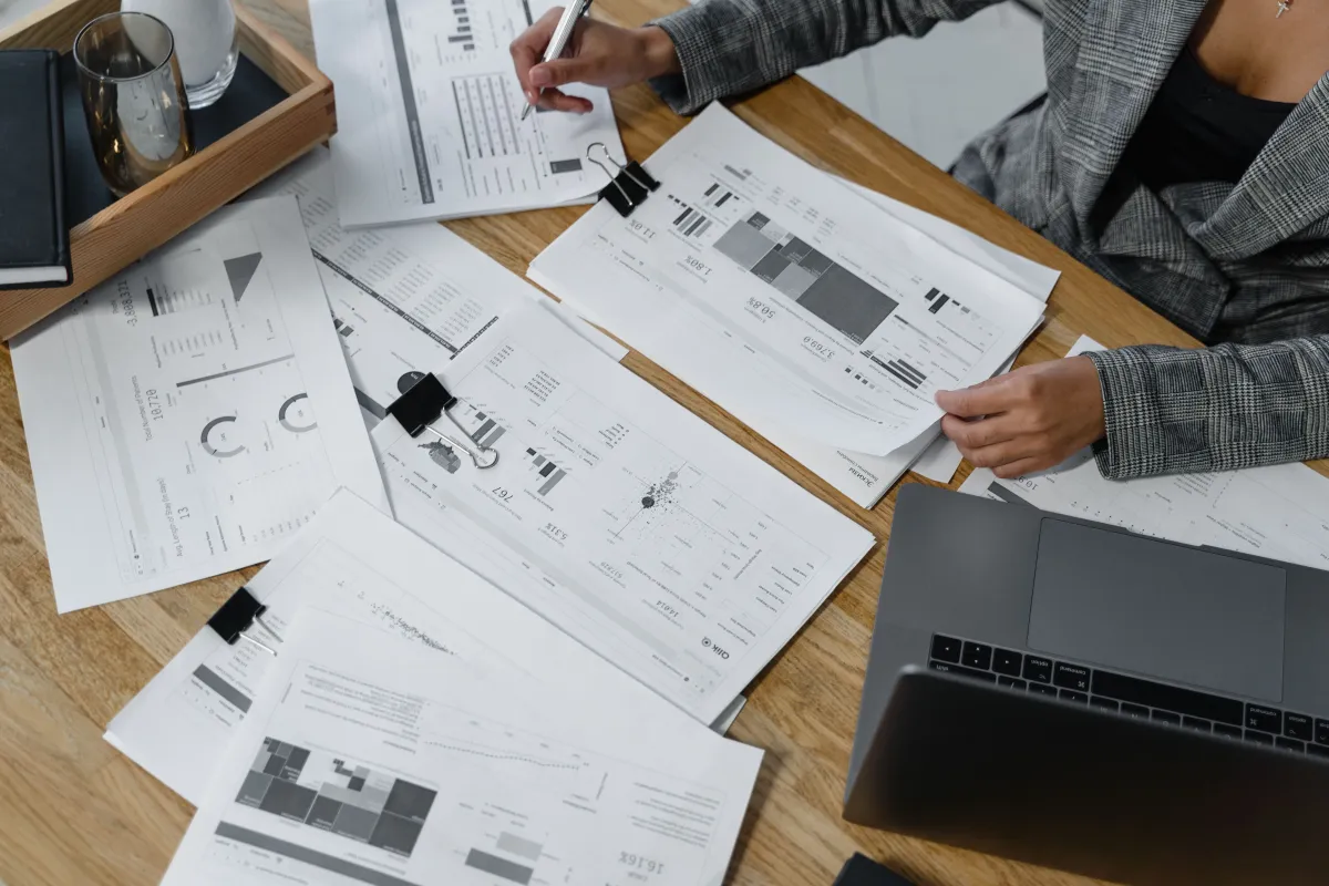Profissional conferindo relatórios impressos e informações financeiras ao lado de um notebook.