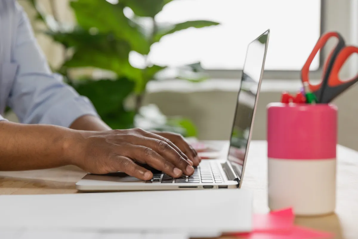 Pessoa utilizando notebook em um escritório, digitando para organizar processos e dados de trabalho; ao fundo há itens de mesa como caneta e organizador, contexto de Sistema ERP.