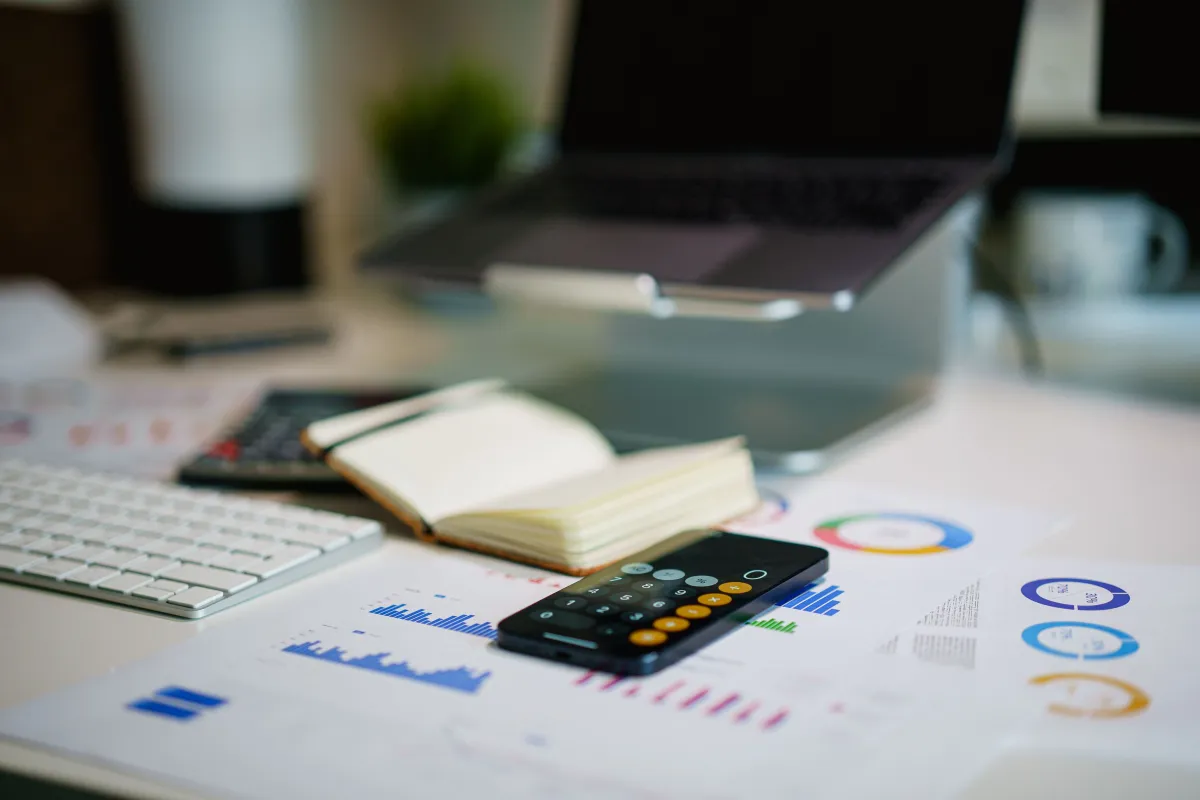 Mesa de escritório com calculadora, notebook aberto e gráficos financeiros coloridos em um relatório, sugerindo organização do Fluxo de Caixa Simples da empresa em poucos passos.