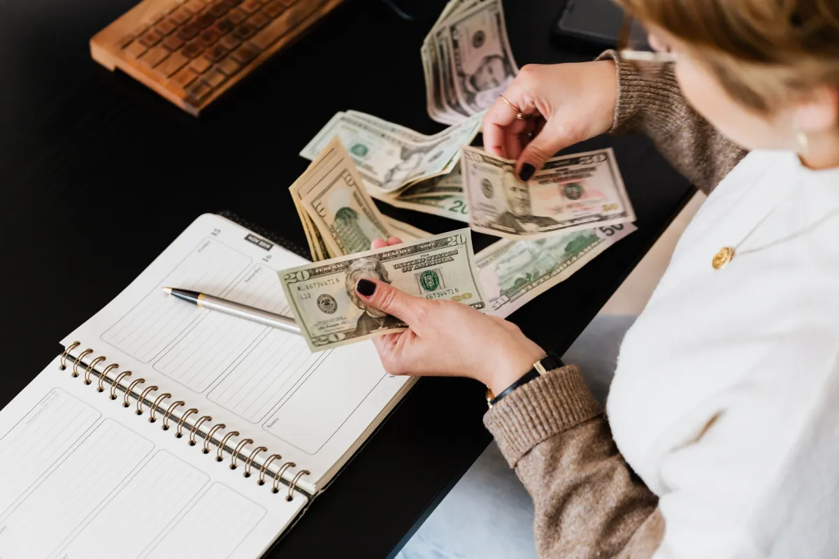 Imagem mostra a organização do financeiro: uma pessoa segurando cédulas de dinheiro enquanto anota valores em um caderno com modelo de planejamento, com foco em Fluxo de Caixa Simp