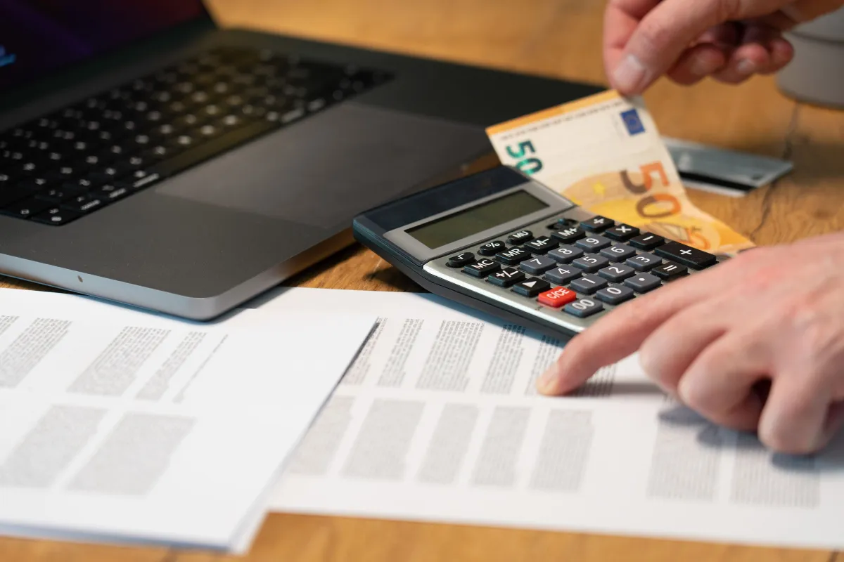 Fluxo de Caixa Simples: mão usando calculadora e contando dinheiro ao lado de notebook e documentos financeiros em uma mesa de trabalho, representando organização do financeiro da