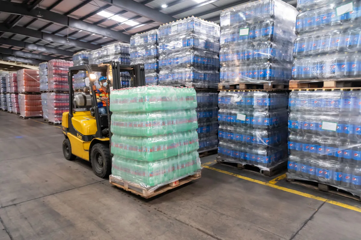 Armazém logístico com empilhadeira carregando pallets com caixas em embalagens plásticas e pacotes empilhados em prateleiras, com galpão e iluminação industrial ao fundo.