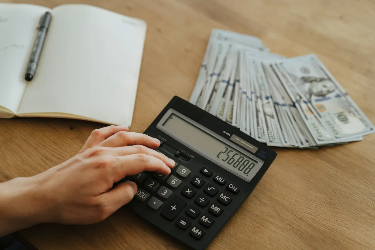 Pessoa calculando valores com calculadora financeira em uma mesa de madeira, com caderno aberto e maço de dinheiro ao fundo