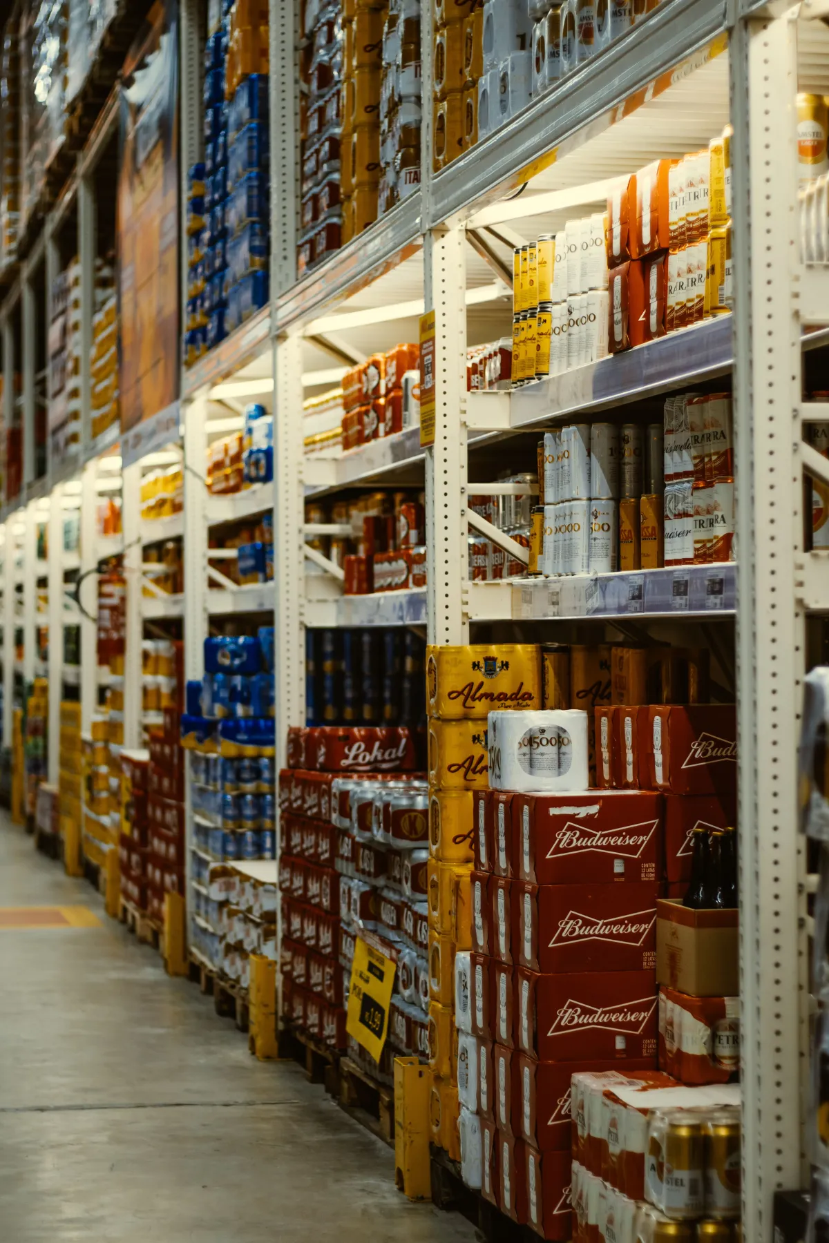 Corredor de supermercado com prateleiras cheias de bebidas alcoólicas enfileiradas em caixas e garrafas, com foco nas marcas de cerveja Budweiser e outras.