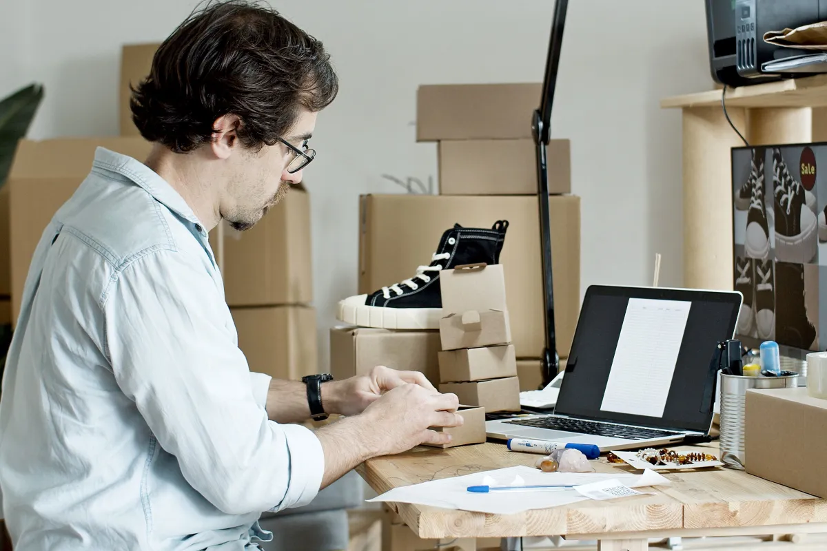 Pequeno empreendedor preparando encomendas sobre uma mesa, com notebook e embalagens.