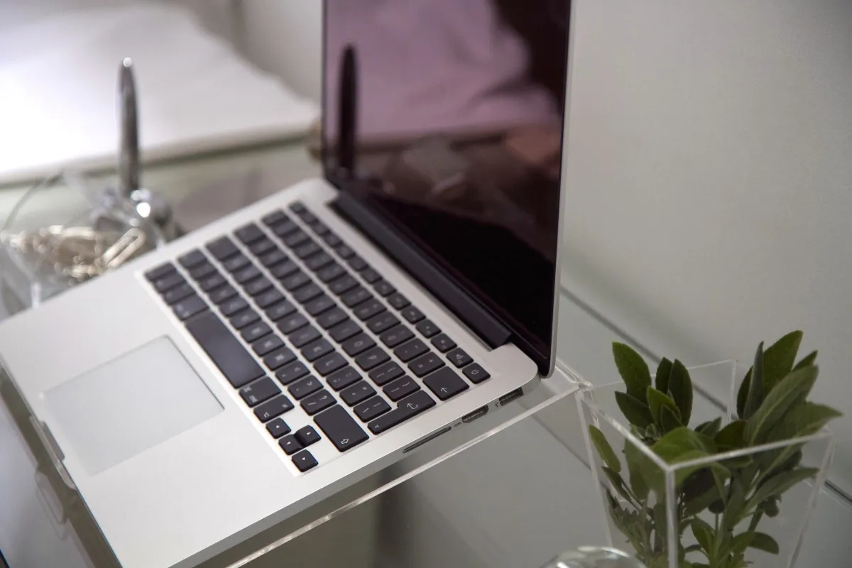 Notebook aberto em uma mesa de trabalho, com foco no teclado e na tela refletindo luz; ao lado há vaso com folhas verdes e objetos decorativos, ambiente corporativo