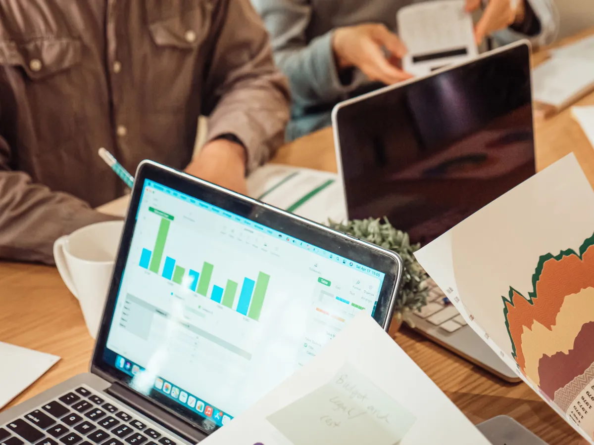 Homem trabalhando em um laptop com gráficos de dados e planilhas na tela, durante uma reunião em escritório, com relatórios impressos e materiais sobre a mesa.